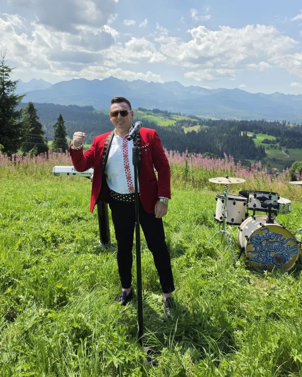 Szalony Drużba w czerwonej marynarce z mikrofonem na hali, w tle panorama Tatr i perkusja z napisem Szalony Drużba Show